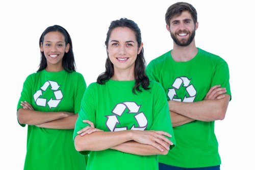A group of three individuals standing outdoors in front of a neutral background, all wearing matching bright green T-shirts featuring a white recycling symbol. The woman in the center has dark hair styled in loose waves, smiling directly at the camera with her arms crossed. To her left, a woman with long dark hair stands with her arms folded, also smiling, while on her right, a man with a beard and short dark hair crosses his arms and smiles. The scene conveys team spirit and environmental awareness associated with rubbish removal and waste management services, aligning with the themes of responsible recycling and sustainability in the context of local Ealing house clearance activities.