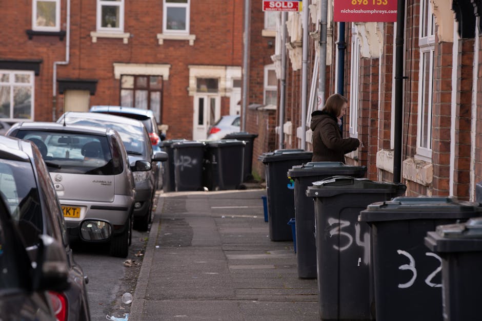 A residential street with a narrow sidewalk lined with multiple black wheelie bins positioned in front of brick terraced houses. The bins are arranged in a line along the curb, with some lids partiall